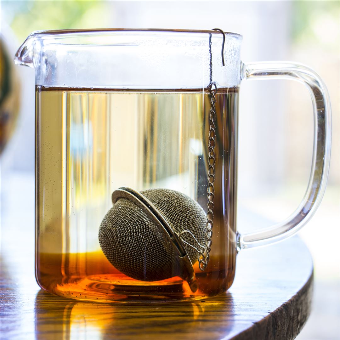 A glass teapot filled with amber tea sits on a wooden table. A Mesh Ball Infuser is submerged in the tea, its chain draped over the rim as sunlight streams in, creating a warm and inviting atmosphere.